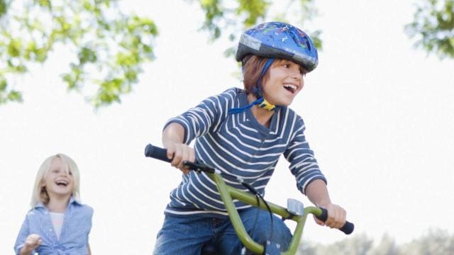 A child wearing a helmet rides a bicycle on grass while another child runs behind, surrounded by greenery and trees.