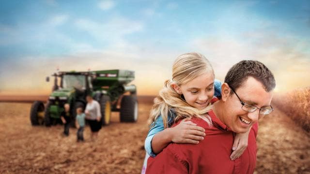 A man gives a piggyback ride to a smiling girl in a field with a tractor in the background.